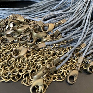 Op de afbeelding: Een stapel grijze koord en goudkleurige metalen sluitingen. Het koord is gemaakt van een dun, geweven materiaal. De sluitingen zijn klein en hebben een kreeftenschaarontwerp.