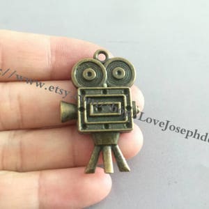 May include: A bronze-colored metal charm shaped like a vintage movie camera. The camera has two round film reels, a rectangular viewfinder, and a small handle on the side. The charm has a loop at the top for attaching to a necklace or bracelet.