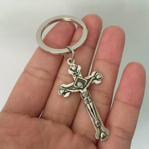May include: Silver-colored crucifix keychain. The cross features a detailed depiction of Jesus Christ and the letters INRI. The keychain has a circular ring for attaching keys. The religious item is held in a hand.