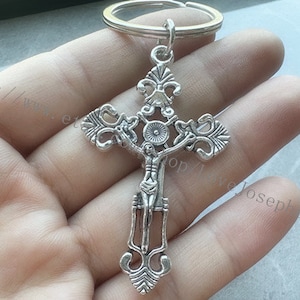 May include: Silver-toned metal cross keychain with a detailed design. The cross features a figure of Jesus Christ on the front. The keychain has a silver-toned metal ring.