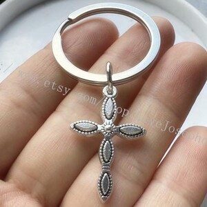 May include: Silver key chain with a silver cross charm. The cross has a detailed design with small oval shapes.
