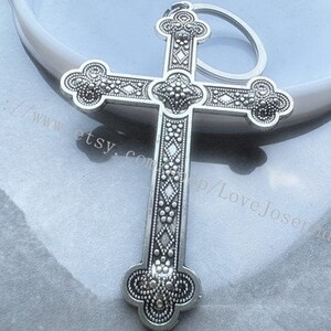 May include: Silver-toned ornate cross keychain with a detailed design. The cross has a diamond pattern and small round accents.