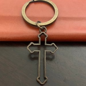 May include: A bronze-colored metal cross keychain with a simple design. The cross is attached to a metal ring.