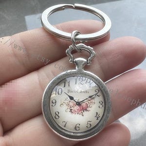 May include: Silver pocket watch keyring with a floral design on the watch face. The watch face has black hands and Roman numerals. The text "Rachel Seeds" is visible. The keyring is attached to a silver ring.