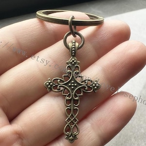 May include: A bronze-colored metal cross keychain with an ornate design. The cross is attached to a silver-colored metal ring.