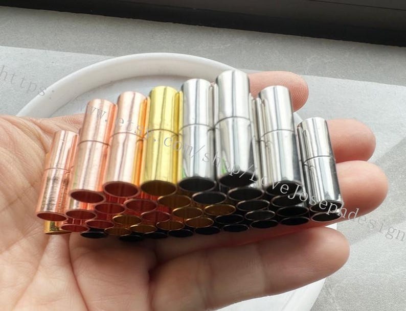 May include: A collection of silver, gold, and rose gold metal end caps for necklaces or bracelets. The caps are cylindrical and have a smooth finish.