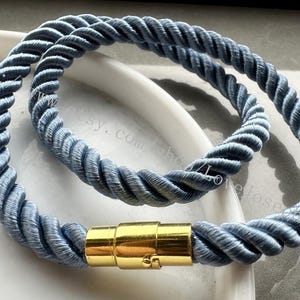 Puede incluir: Un par de pulseras de cuerda azul con un cierre magnético dorado. Las pulseras están hechas de una cuerda gruesa trenzada y están diseñadas para usarse en la muñeca.