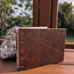 May include: A rectangular journal with a dark brown, textured leather cover. The spine features exposed stitching with thin, light-colored thread that binds the white, rough-edged paper pages together in a horizontal, landscape orientation.