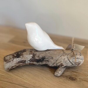 Könnte beinhalten: Eine glatte, weiße Keramikvogel-Figur, die auf einem Stück verwittertem, strukturiertem Treibholz sitzt. Der Vogel hat eine einfache, abgerundete Form ohne sichtbare Gesichtszüge. Ein kleines Stück Bindfaden ist um das Holz nahe dem Schwanz gebunden.