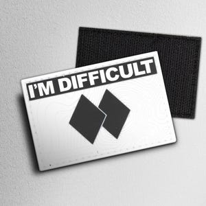 May include: A white patch with the text "I'M DIFFICULT" in black, above two black diamond shapes. The patch has a topographic map design and a black hook-and-loop backing.