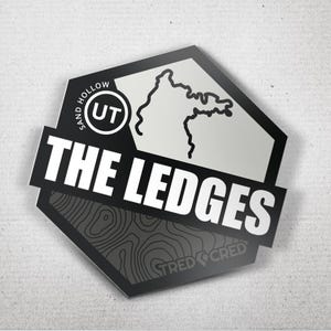 May include: A black and white sticker with the text "THE LEDGES" in bold white letters. The sticker has a hexagonal shape with a map outline and the text "SAND HOLLOW UT". The bottom section has a topographic map design and the text "STRED CRED".