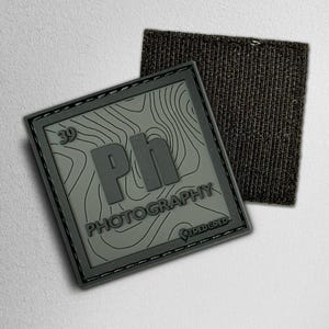 May include: A gray square patch with the word "PHOTOGRAPHY" and the letters "Ph" in a stylized design. The patch has a topographic map pattern and the number "39". A separate black fabric square is next to the patch.