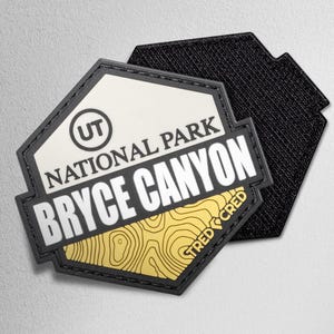 May include: A black and white Bryce Canyon National Park patch with a topographic map design in gold. The patch features the text "NATIONAL PARK" and "BRYCE CANYON" in white, with "UT" in a circle. The patch is hexagonal.