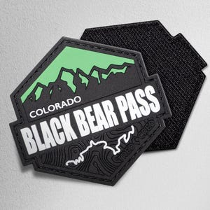 May include: Black and green hexagonal patch with the text "COLORADO BLACK BEAR PASS" in white. The patch features a mountain range graphic in green and black, and a topographic map design. The back of the patch is black.