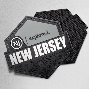 May include: A black and gray patch with the text "NEW JERSEY" in white block letters. The patch has a topographic design and the words "explored." and "NJ" are also visible. The patch is a hexagon shape.