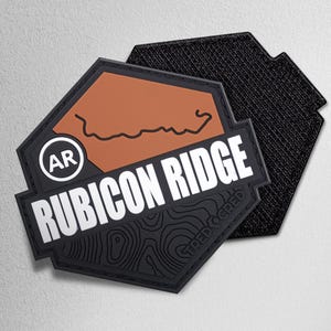 May include: A black and brown patch with the words "RUBICON RIDGE" in white. The patch has a topographic design and the letters "AR" in a white circle. The patch is hexagonal and has a hook and loop backing.