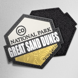 May include: A black and white patch with the text "NATIONAL PARK GREAT SAND DUNES" and a topographic map design. The patch also features the letters "CO" inside a circle. The patch is hexagonal in shape and has a black velcro backing.