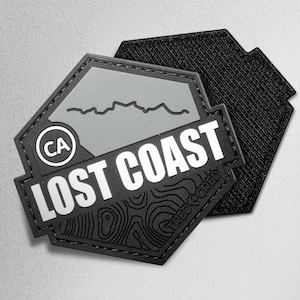May include: A black and gray patch with the words "LOST COAST" in white. The patch has a hexagonal shape with a gray background and a mountain range graphic. The patch also features the letters "CA" in a circle and the words "TRED CRED".