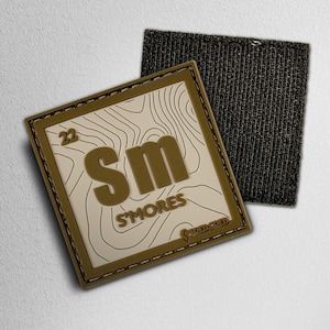 May include: Square patch with a topographic map design and the text "Sm'ores". The patch has a brown border and a hook-and-loop backing. The patch also has the text "TIRED CRED".
