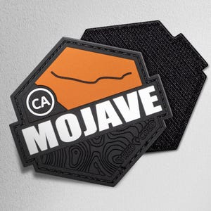 May include: A black and orange Mojave patch with white text. The patch has a hexagon shape with a black border. The text "MOJAVE" is in bold white letters. The patch also features the letters "CA" inside a white circle.