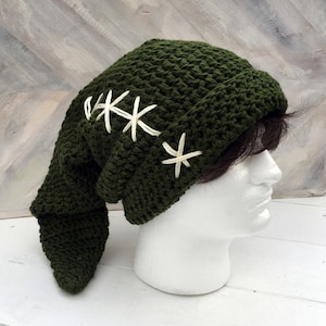 May include: A dark green, crocheted hat with a long, pointed flap. The hat features three white, star-shaped embroidered accents. The hat is displayed on a white mannequin head.