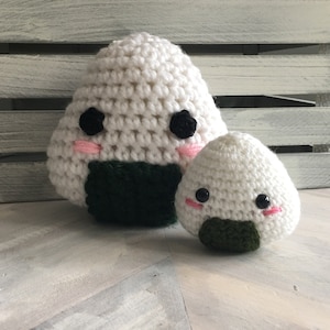 May include: Two crocheted onigiri, a Japanese rice ball, with cute faces. The larger onigiri is white with black eyes and pink cheeks, and the smaller onigiri is white with black eyes, pink cheeks, and a green seaweed wrap.