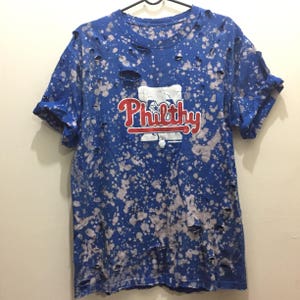May include: A blue t-shirt with white bleach splatters and holes. The shirt has a red and white logo that says "Philthy" on the front.