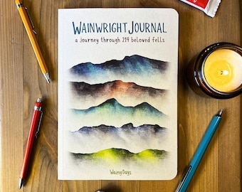 May include: A cream-colored journal titled Wainwright Journal: a Journey through 214 beloved fells, featuring a multi-layered mountain illustration in blue, brown, green, and purple hues, accompanied by four pens in yellow, red, and blue.