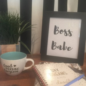 Puede incluir: Una taza de cerámica blanca con interior turquesa y el texto "Good Morning Gorgeous" se encuentra junto a una impresión enmarcada que dice "Boss Babe". Un cuaderno y un bolígrafo también están sobre la superficie de madera, con una planta en una maceta de madera en el fondo.