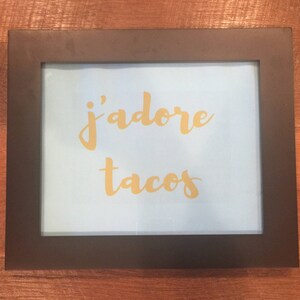 May include: A framed print with a light blue background and gold lettering that says "j'adore tacos".