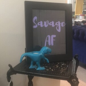 Peut inclure: Une image encadrée noire avec les mots "Savage AF" en lettres violettes. Une figurine de dinosaure bleue est posée sur une table en métal noir.