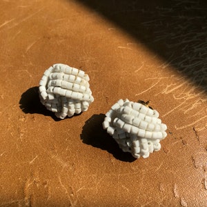May include: A pair of white beaded earrings, each shaped like a knot. The earrings are made of small, square beads that are strung together to create a three-dimensional design.