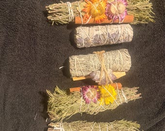 Puede incluir: Cinco manojos de hierbas y flores secas dispuestos en fila vertical. Tres manojos incluyen romero, ramas de canela y flores secas coloridas. Un manojo está envuelto en hilo blanco y otro incluye un pequeño cristal de color morado.