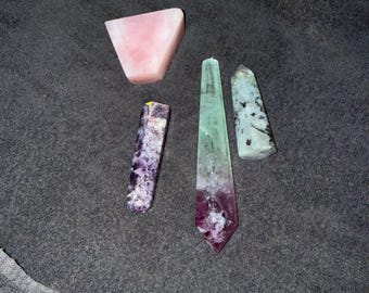 Puede incluir: Una colección de cuatro piedras pulidas que incluye una pieza triangular de color rosa claro, una punta de cristal rectangular de color morado y blanco, una torre de cristal alta y puntiaguda de color verde y morado, y una piedra cilíndrica moteada.