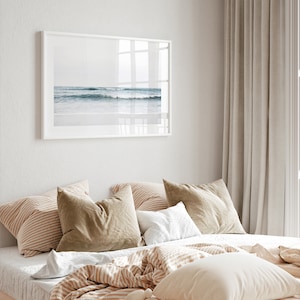 May include: A bedroom with a white framed picture of a beach scene above a bed with a white, brown, and beige striped comforter and pillows. The curtains are a light beige color.