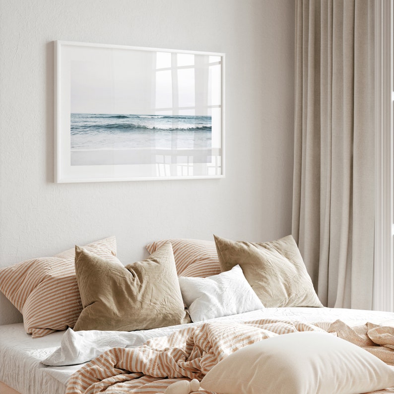 May include: A bedroom with a white framed picture of a beach scene above a bed with a white, brown, and beige striped comforter and pillows. The curtains are a light beige color.