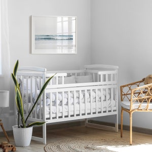 May include: White wooden crib with a white mattress and white bedding. The crib is in a white room with a wicker chair and a framed picture of a beach scene.