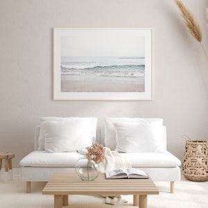 May include: A white couch with two white pillows and a throw blanket sits in front of a framed photograph of a beach scene. A vase with dried flowers sits on a wooden coffee table in front of the couch.