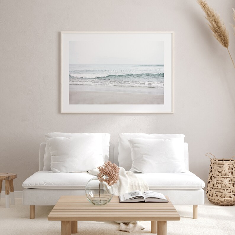 May include: A white couch with two white pillows and a throw blanket sits in front of a framed photograph of a beach scene. A vase with dried flowers sits on a wooden coffee table in front of the couch.