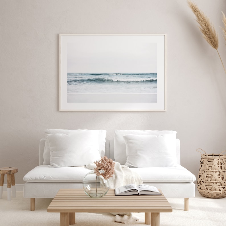 May include: A white couch with two white pillows and a throw blanket sits in front of a white wall with a framed photograph of ocean waves. A small wooden stool and a woven basket are on the floor. A vase with pink flowers sits on a wooden coffee table.