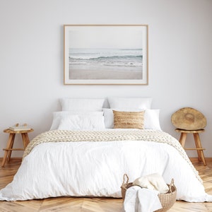 May include: A white bed with a white duvet cover and a knitted throw blanket. The bed has two white pillows and a tan pillow. There is a framed photograph of a beach scene above the bed. There are two wooden side tables with books on them.