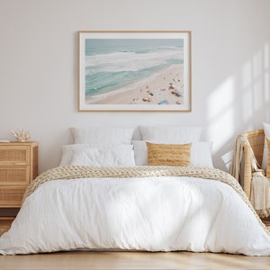 May include: A white bed with a white duvet cover and a tan knitted throw blanket. The bed has two white pillows and a tan pillow. A framed photograph of a beach scene hangs above the bed.