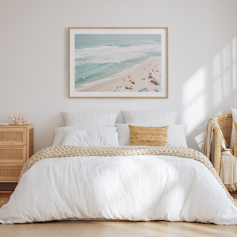 May include: A white bed with a white duvet cover and a tan knitted throw blanket. The bed has two white pillows and a tan pillow. A framed photograph of a beach scene hangs above the bed.