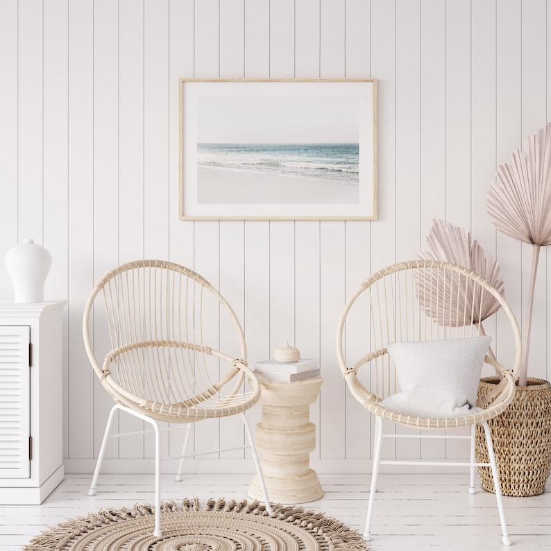 May include: Two white wicker chairs with white metal legs, a small wooden side table, and a framed photograph of a beach scene. The chairs are set against a white wood panel wall.
