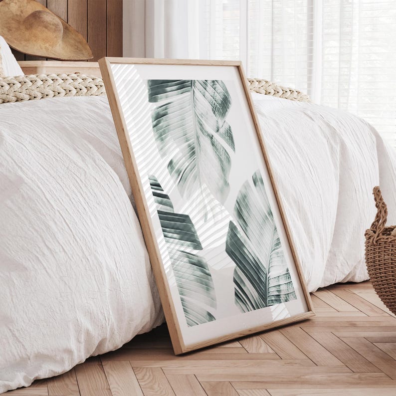 May include: A framed art print featuring a botanical design of large, stylized green and white leaves. The artwork is displayed in a light wood frame and leans against a white bed with a woven headboard. The room has a bright, airy feel.