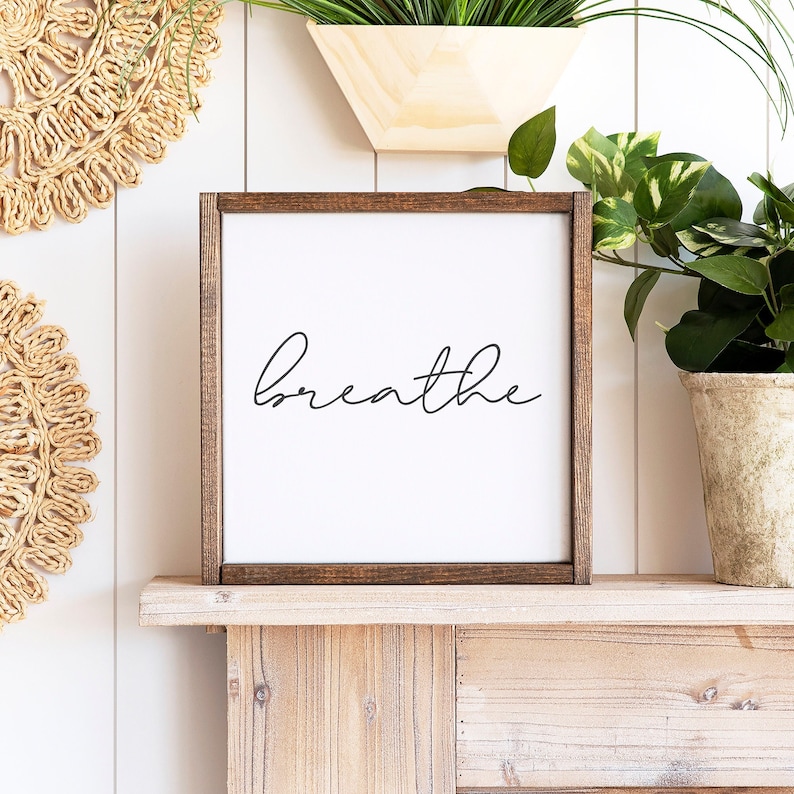 May include: Square wooden framed sign with the word "breathe" in black script on a white background. The sign is displayed on a wooden shelf with plants and decorative elements in the background.
