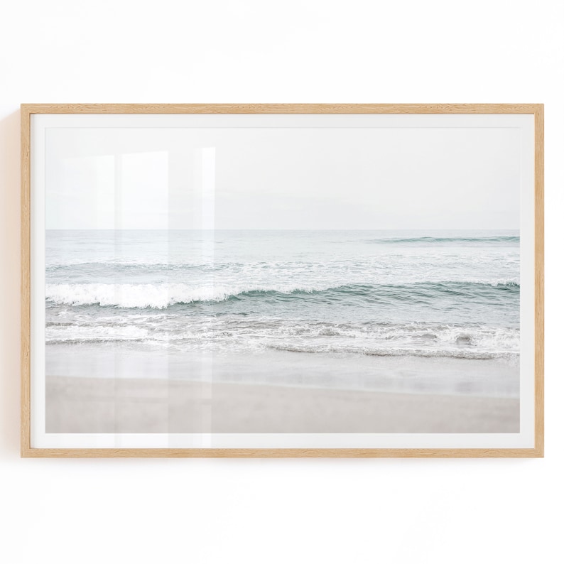 May include: A framed photograph of a beach scene with waves crashing on the shore. The water is a light blue and green color, and the sand is white. The sky is a light gray color.