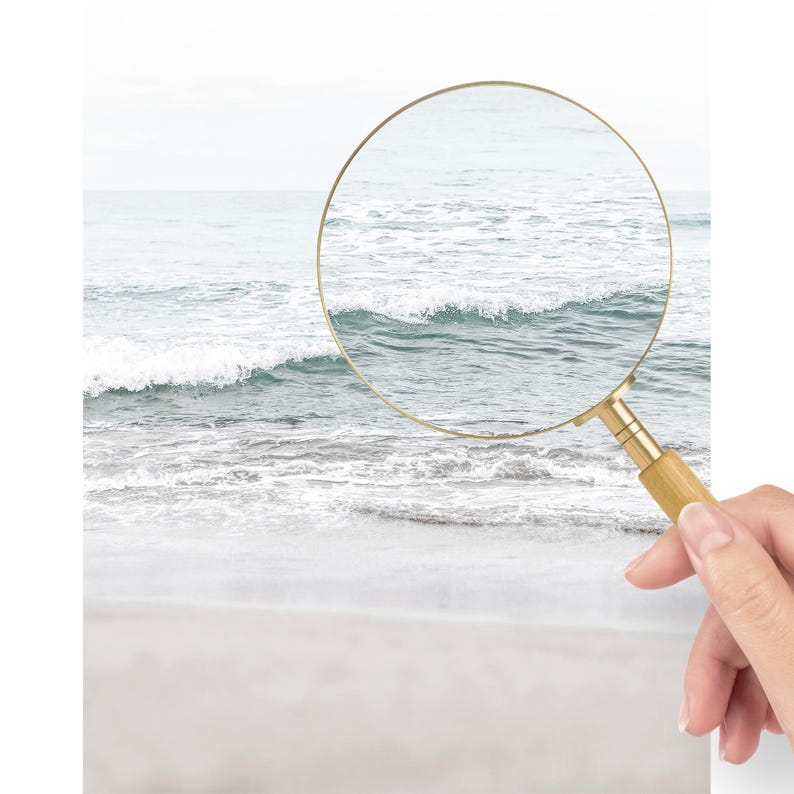May include: A magnifying glass is positioned over a photo of a beach scene. The glass is focused on a wave breaking on the shore. The water is a light blue color and the sand is white.