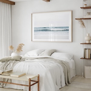 May include: A white bed with a gray blanket and a framed photograph of a beach scene above it. The photograph shows a blue ocean with waves crashing on the shore.