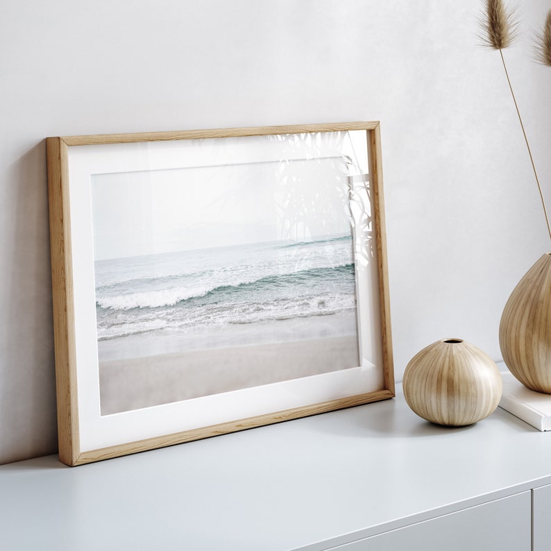 May include: A framed photograph of a beach scene with waves crashing on the shore. The frame is made of light wood and has a white mat.